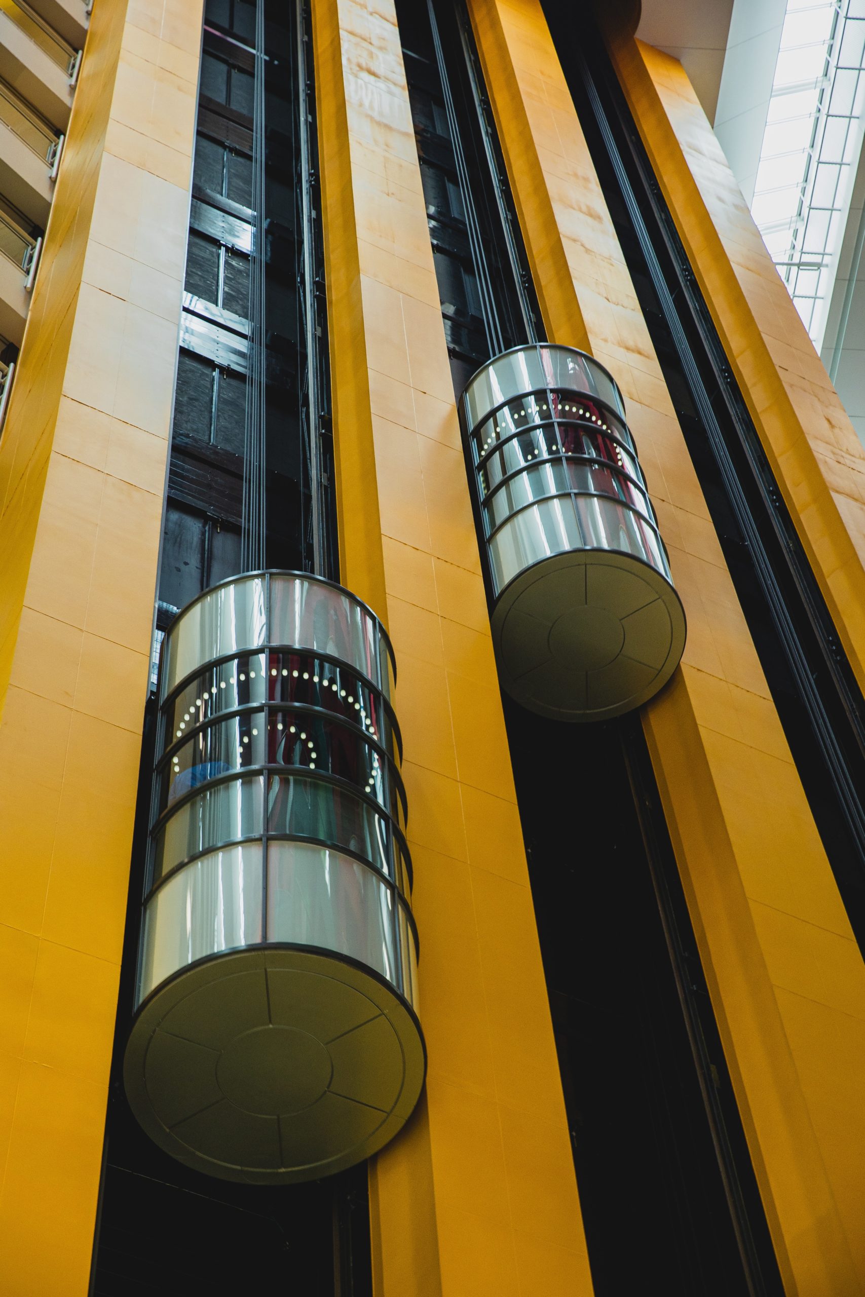 Color photograph of the outside of a tall building. The building has orange colored metal columns reaching upwards. Between columns are tubelike elevators - outside the building running along the face.