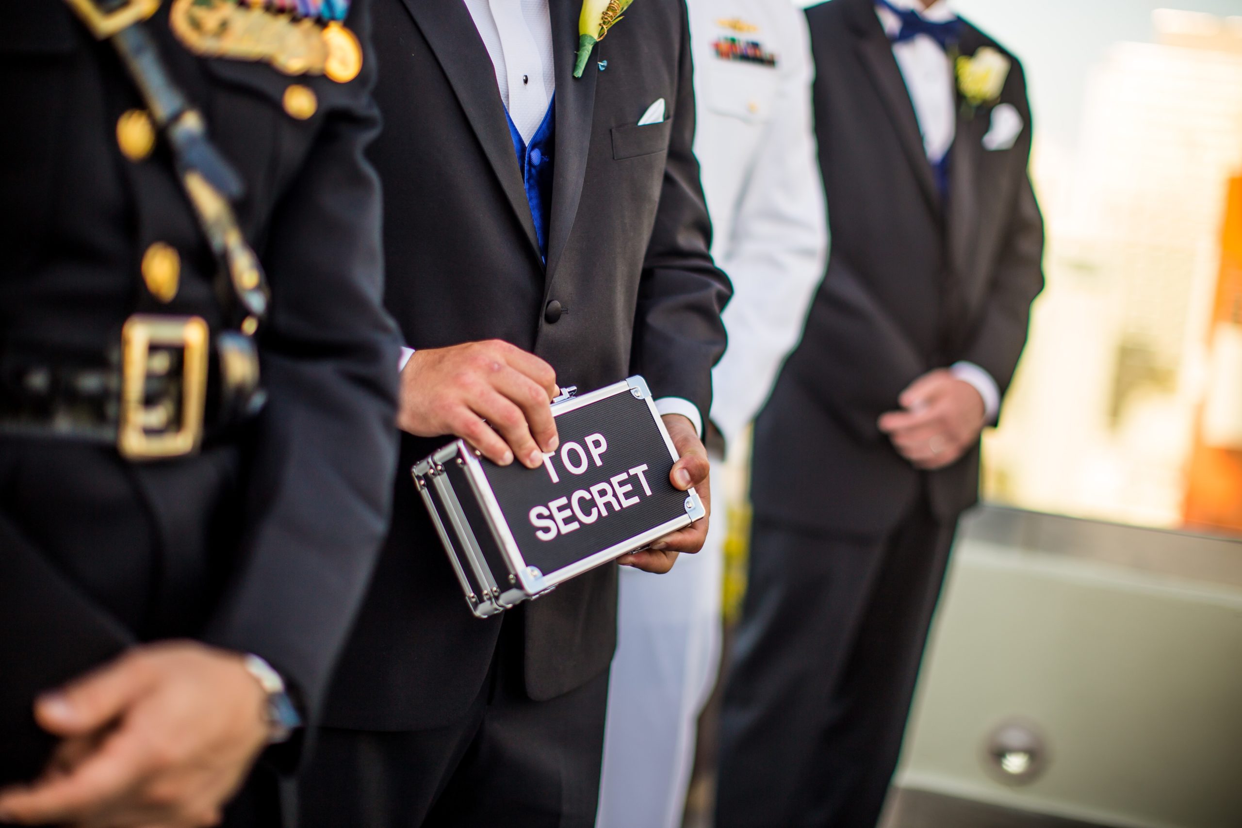Several uniformed men standing next to each other. Faces are not seen. One holds a small case with the words "Top Secret" on the side.